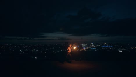 young blond male does tricks with fire breaths fire in the middle of the night with city skyline in background