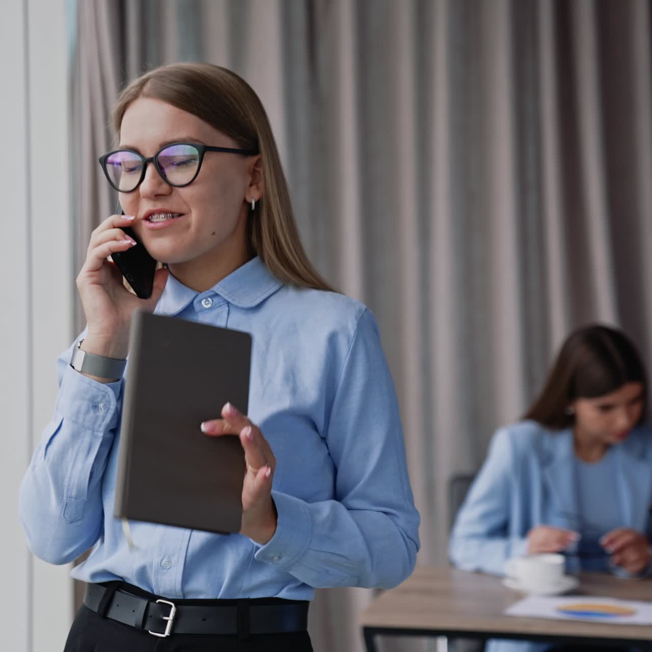 Happy smiling female in glasses speaks on the phone holding notebook in her hand. Colleagues communicate looking at man's phone