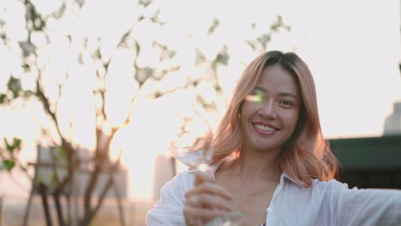 Wanita Muda Ceria Memegang Gelas Anggur Saat Matahari Terbenam di Rooftop