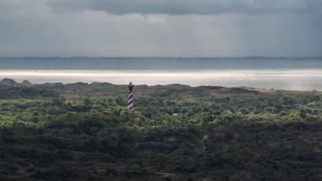 Vista-Aérea-En-órbita-De-Una-Torre-De-Faro-De-Rayas-Rojas-Y-Blancas-Que-Sobresale-De-Los-árboles-Con-Vistas-A-La-Costa-Con-Lluvia-Y-Nubes-Rodando-Por