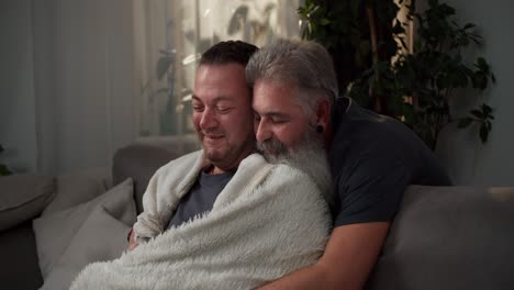 happy brunette man with stubble in a white woolen blanket drinks hot tea and hugs with his man a guy with gray hair and a lush gray beard on a cozy evening in a modern apartment sitting on the sofa