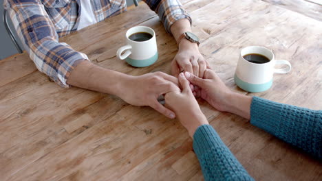 midsection of biracial couple sitting at table, drinking coffee, holding hands at home, slow motion