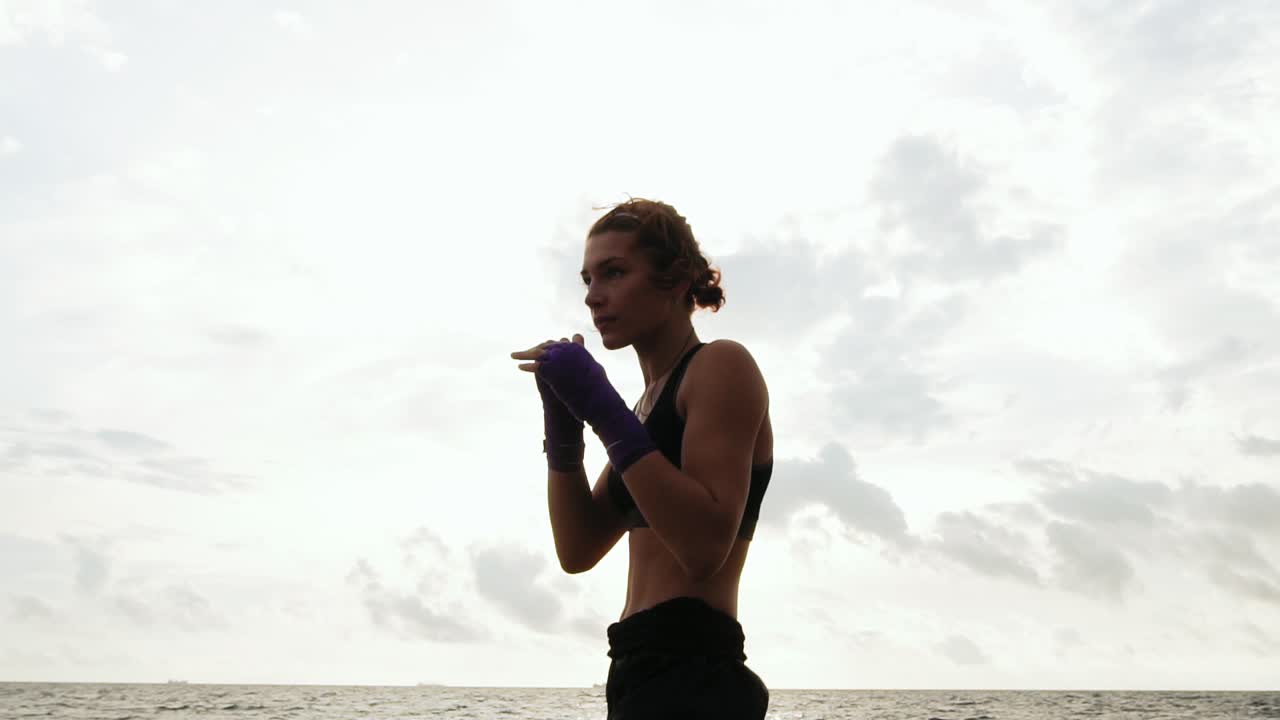 mujer dura y bonita boxeando en la sombra. hermosa boxeadora entrenando junto al mar en la playa por la mañana, lanzando golpes en