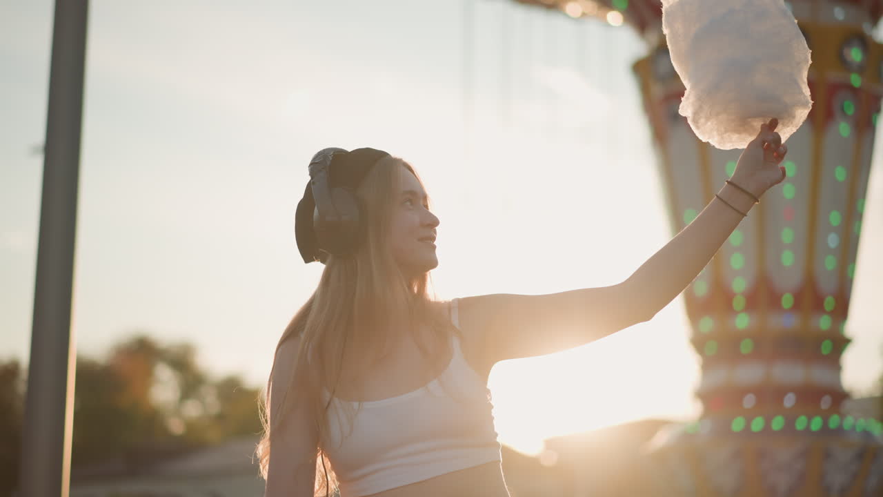 Mulher branca pegando algodão doce sob a luz quente de um parque de diversões, braço estendido e fones de ouvido, momento divertido de frequentadora de festival com brinquedo iluminado atrás, iluminação periférica do sol e gesto espontâneo