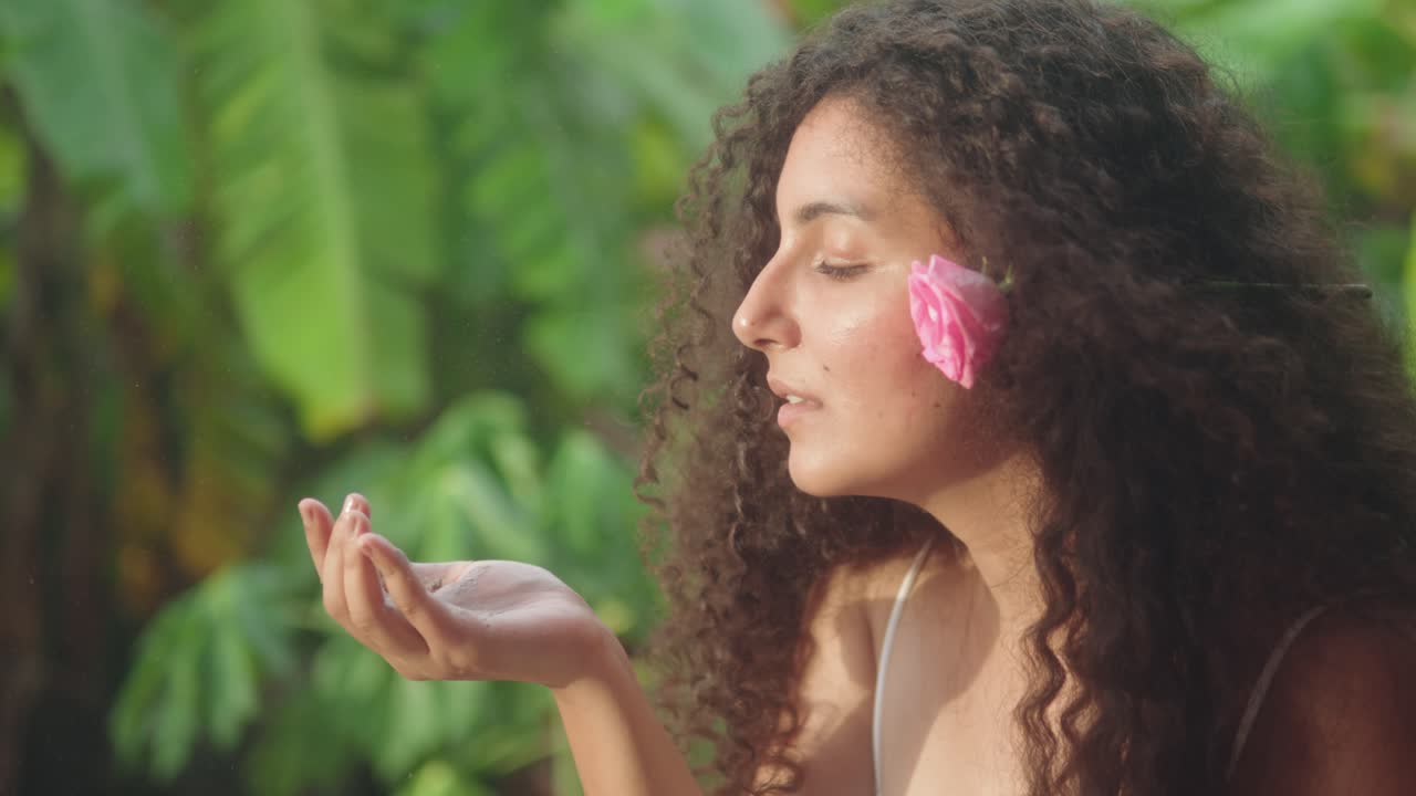 Close-up captures a woman with a pink flower tucked behind her ear, gently blowing ceremonial powder from her hand in a lush green setting, suggesting a ritual or symbolic gesture