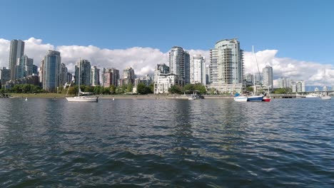 yachts cataramarans sail boats parked on the ocean without a dock next to a luxury park with modern condominium towers that are reflecting the calm water on a sunny summer day with a lush blue sky