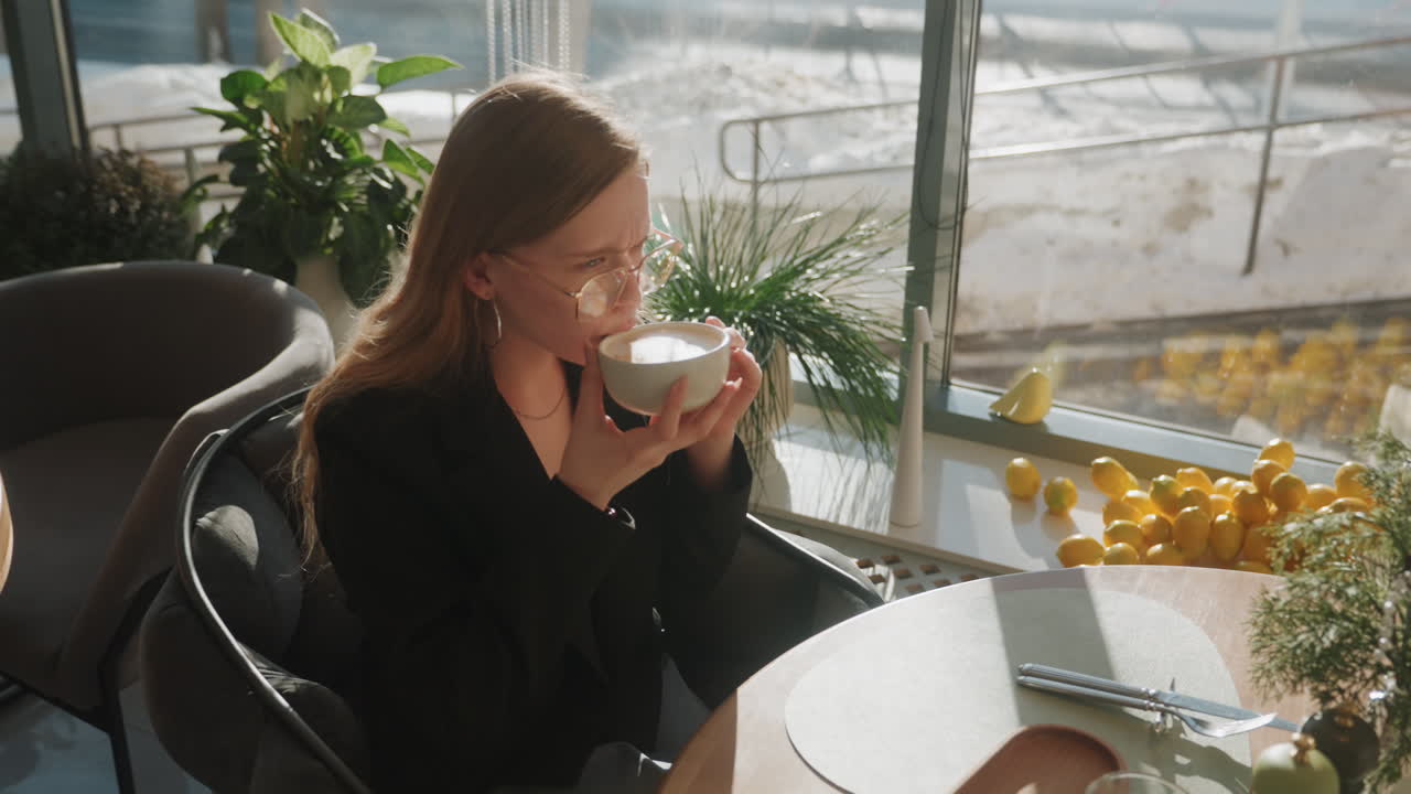Mulher caucasiana em ritual de café divertido, ajustando os óculos e sorrindo enquanto toma de uma xícara de cerâmica, fundo ensolarado com exposição de limões e plantas, blazer chique e vibração de estilo de vida de moda espontânea.