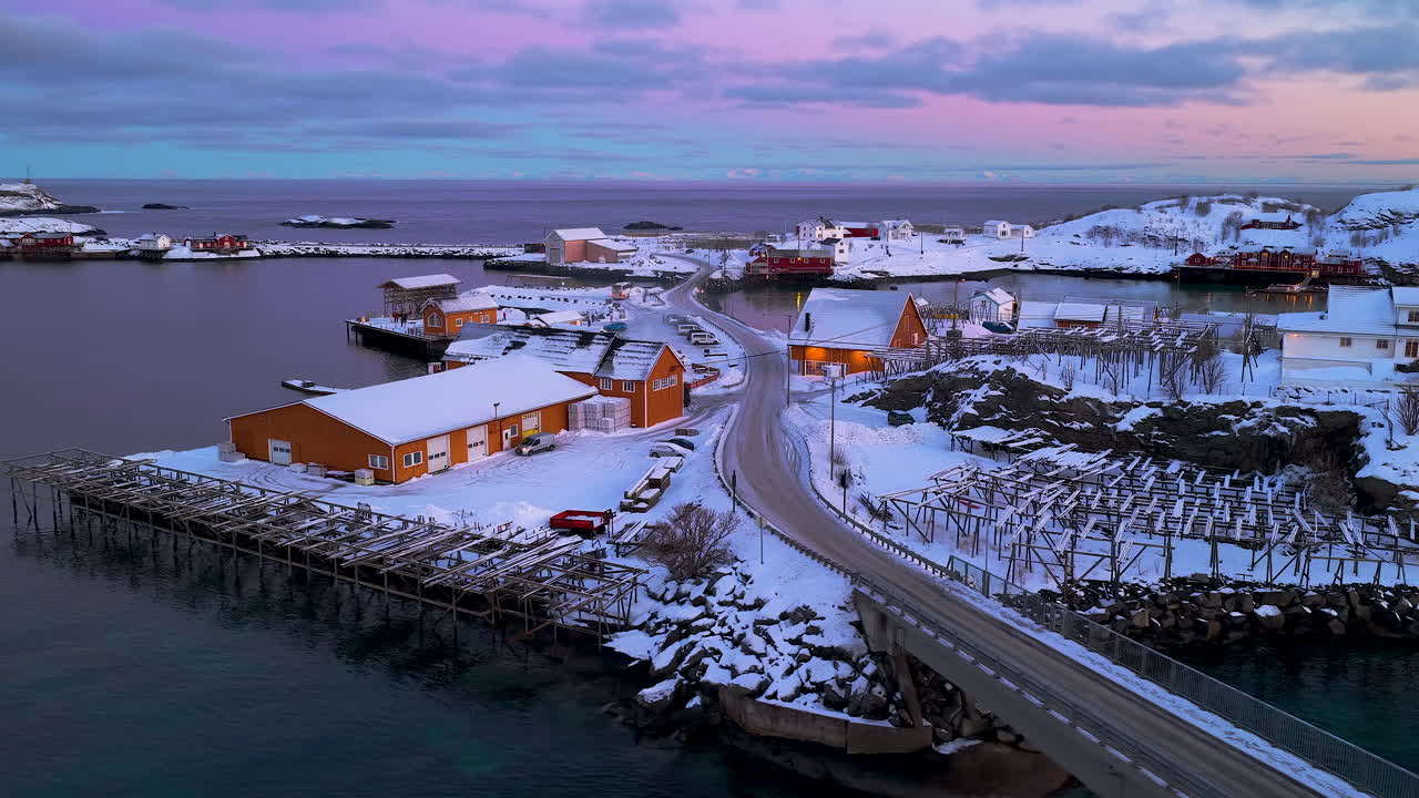 Filmische Drohnenaufnahme eines schneebedeckten norwegischen Fischerdorfes auf den Lofoten bei Sonnenuntergang. Mit traditionellen Rorbu-Hütten und Fischtrockengestellen.