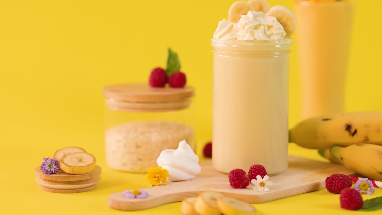 Una mano aggiunge fette di banana a un frullato cremoso di banane, circondato da frutta su sfondo giallo brillante