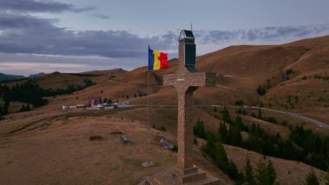 aerial orbiting shot around cross of dichiu mountain with waving romanian flag during cloudy day on summit