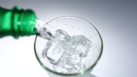 pouring sparkling soda water into glass with ice cubes from green bottle