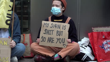 a masked protestor wearing an orange beanie and dungarees sits crosslegged holding a cardboard placard that reads, “my sign is shit but so are you”.