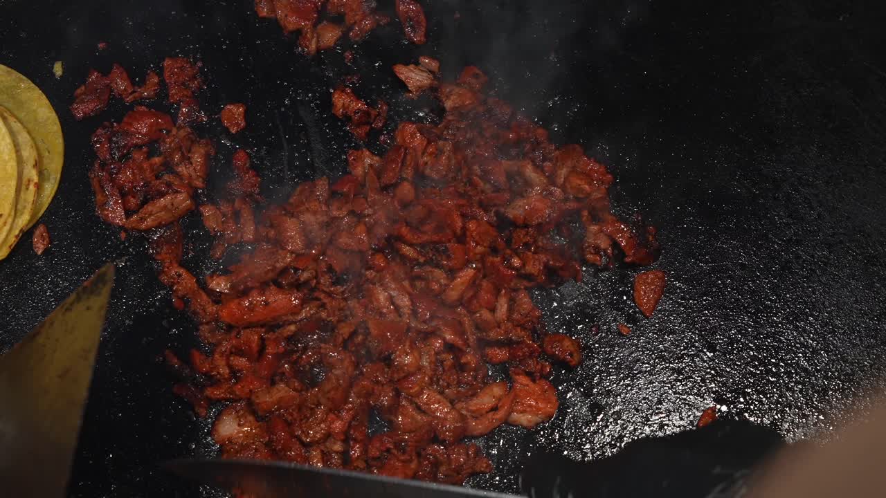 Primo piano POV di carne al pastor sfrigolante in cottura e manipolata con una spatola e un coltello grande su una griglia da street food messicano, con i tacos a lato, che mette in risalto sapore, vapore e aroma salato.