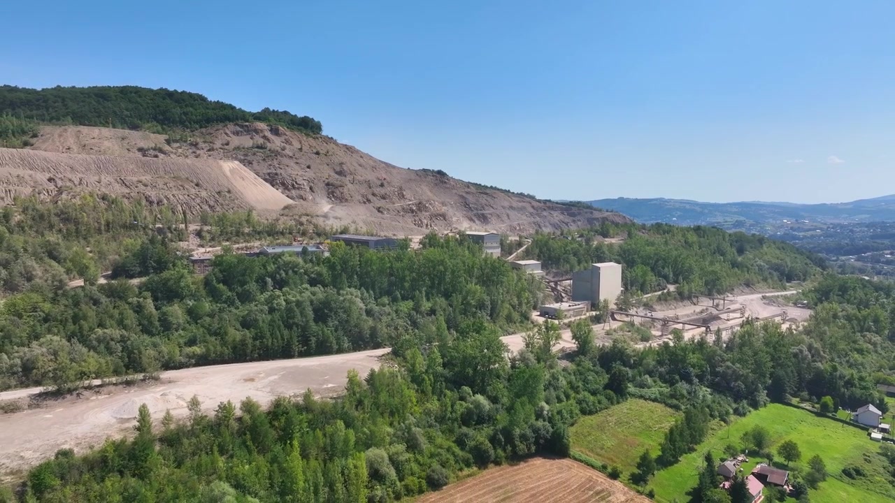 Vista aerea di un sito di cava industriale con edifici, strade e la circostante foresta verde in terreno collinare, ripresa da drone.