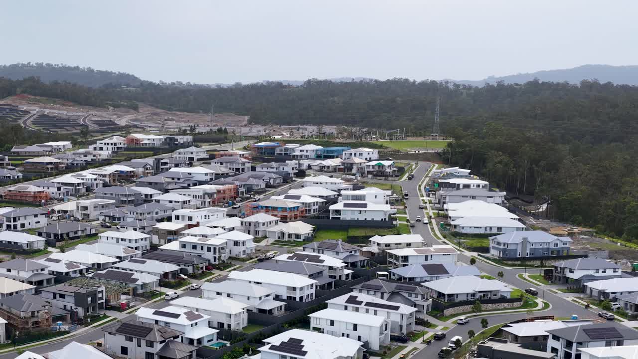 Drone glides over contemporary suburban homes, forest edge, and winding streets under overcast daylight
