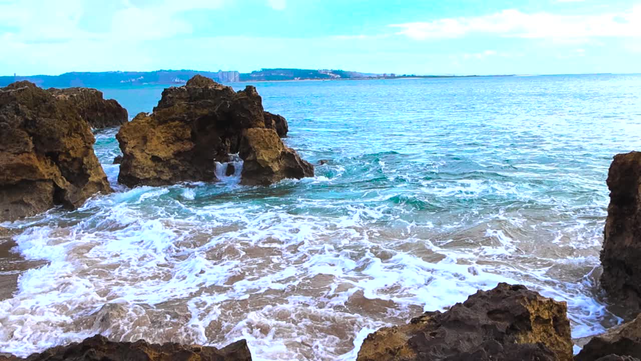 Slow motion handheld footage of foamy water striking sharp rocks and sand along the Caxias shoreline in Portugal with distant hills on the horizon