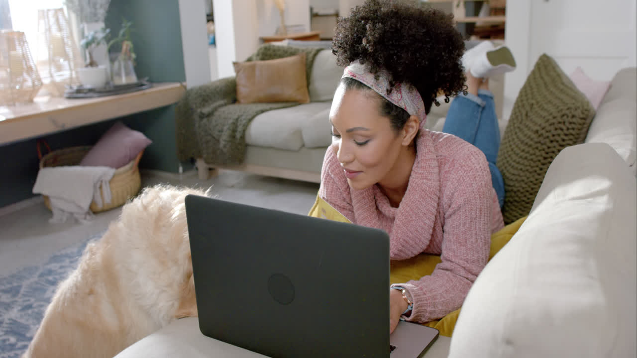 Happy biracial woman with golden retriever dog using laptop and credit card at home, slow motion