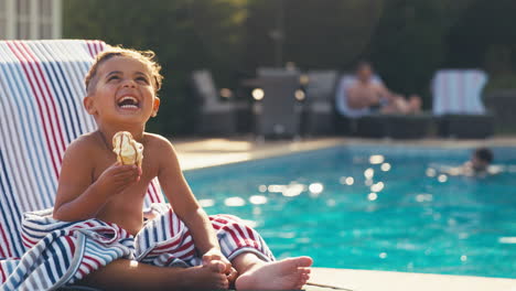 Lachender-Junge-Im-Sommerurlaub-Auf-Der-Liege-Am-Schwimmbad,-Der-Eis-Isst