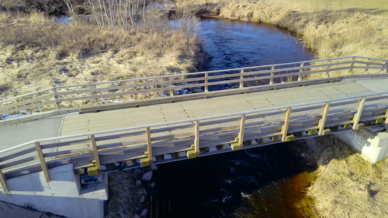 Aerial circling shot of wooden bridge over flowing river. Riverbanks are lined with dried weathered grass. Empty footbridge overcrossing river rapids during sunny day. Open air natural environment
