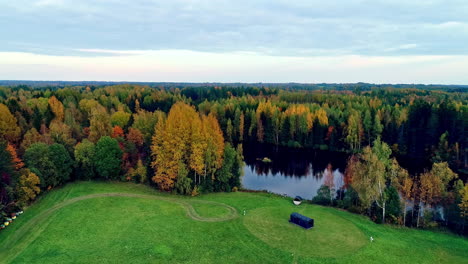 Drone-overview-shot-over-rectangular-vacation-cabin-on-the-bank-of-a-lake-surrounded-by-autumnal-trees-in-a-scenic-rural-countryside-on-a-cloudy-day