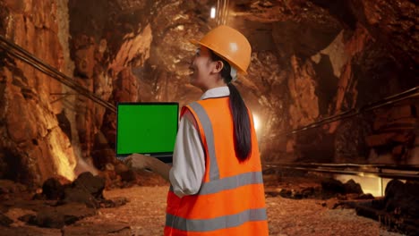 back view of asian female engineer with safety helmet working on a green screen laptop and looking around in underground mine tunnel