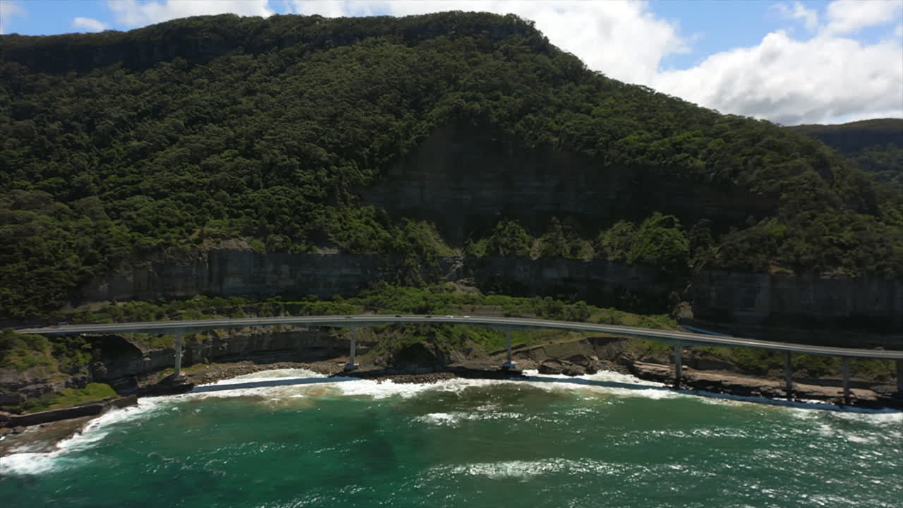 Aerial panning view over Pacific Ocean of Sea Cliff Bridge curving around scenic coastline in Australia.