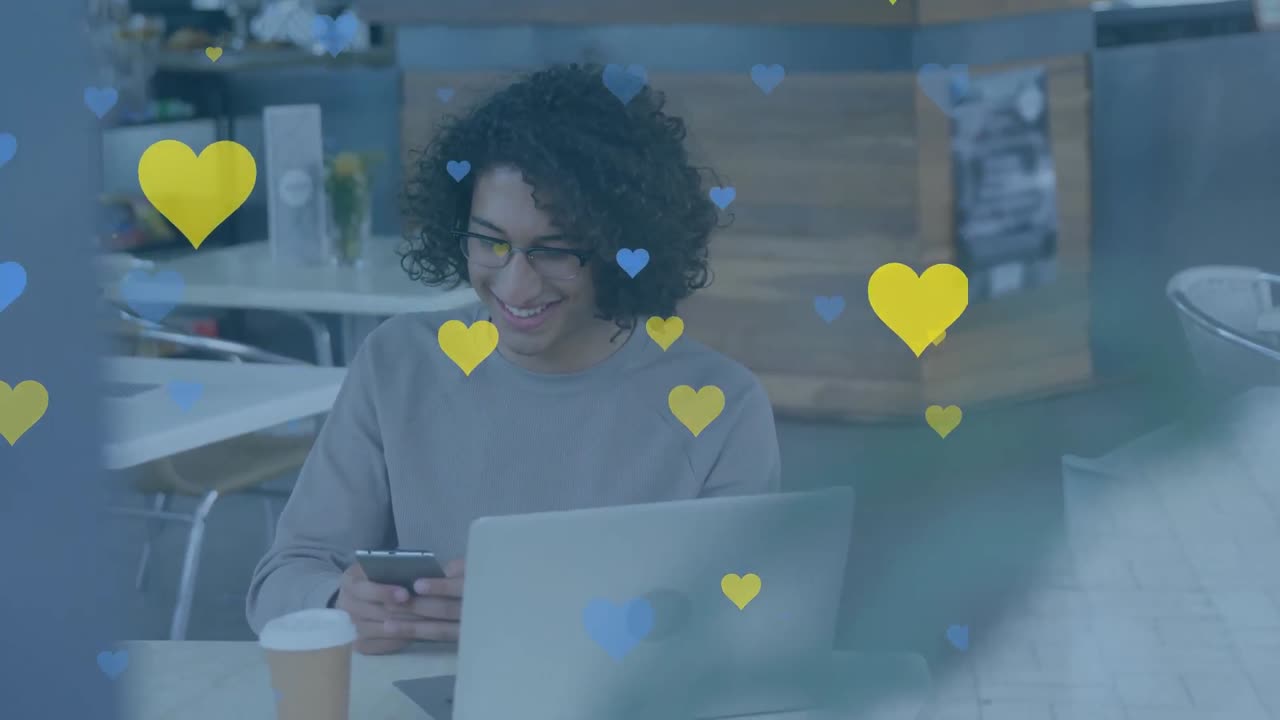 animación de iconos de corazón amarillo y azul sobre un hombre biracial usando un teléfono inteligente en una cafetería
