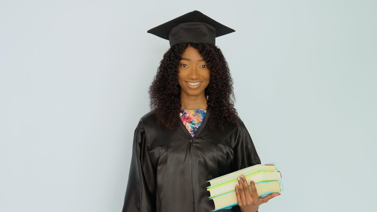 A young African-American woman in a black robe and a master's hat stands straight holding textbooks and looks at the camera. Portrait of half height