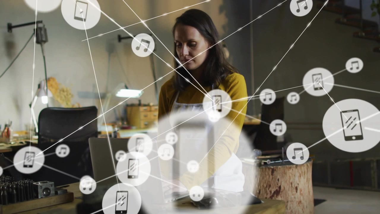 Mid-adult woman typing on laptop in studio visualizing tech network with smartphone and music icons