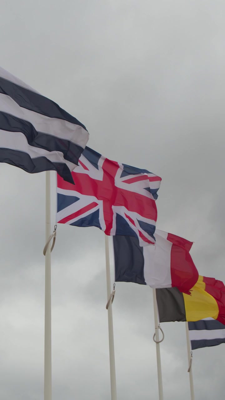 Multiple national flags wave vigorously on tall poles under cloudy skies at coastal landmark