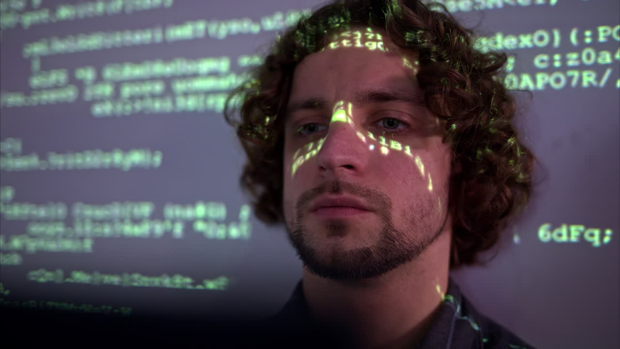 Man with curly hair in front of a projection of green computer code, illuminating on his face