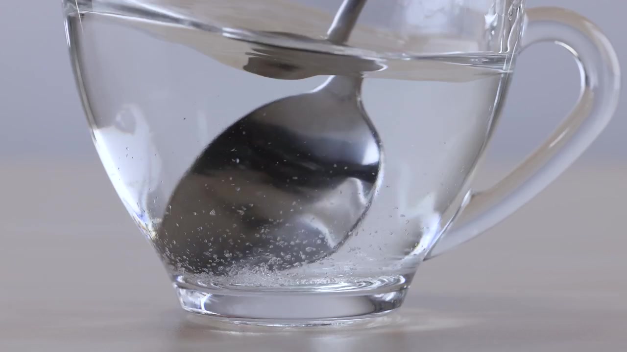 Un cucchiaio di metallo mescola cristalli di zucchero bianco in acqua calda all'interno di una tazza di vetro trasparente