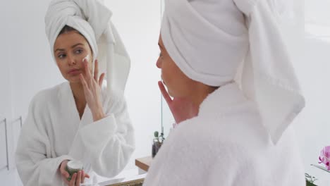 woman in bathrobe applying face cream while looking in the mirror