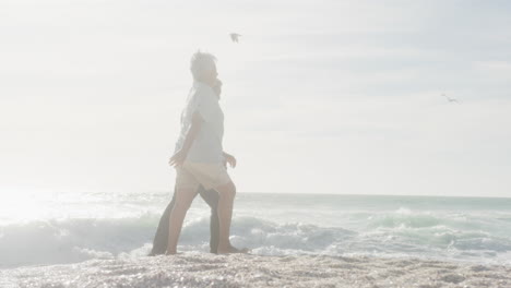 Side-view-of-happy-hispanic-senior-couple-holding-hands,-walking-on-beach-at-sunset