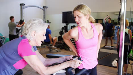 Mujer-Mayor-Haciendo-Ejercicio-En-Una-Máquina-De-Ciclismo-Siendo-Alentada-Por-Un-Entrenador-Personal-En-El-Gimnasio