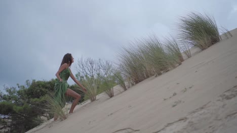mujeres de cabello oscuro con un vestido elegante calssy verde smaragd caminan hacia arriba de una colina de dunas de arena con un cielo gris azulado nublado - cámara lenta cinematográfica dramática