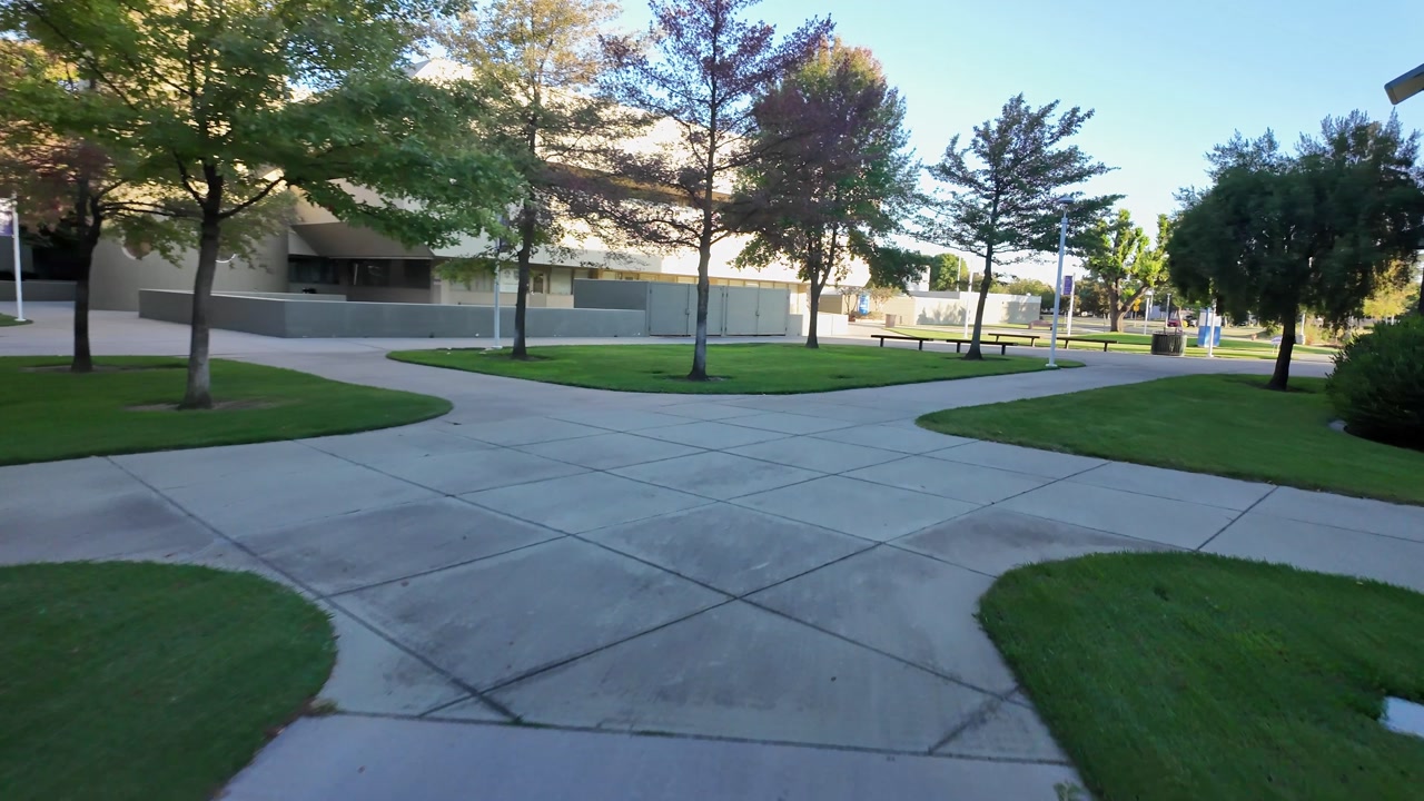 Point of view video walking along a concrete path through the lush green grounds of Modesto Junior College in California, surrounded by trees and modern buildings on a sunny day