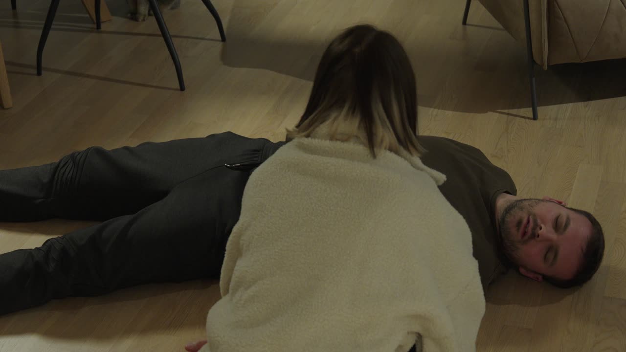 Primo piano di una donna che valuta la respirazione di un uomo incosciente a terra, dimostrando prontezza di risposta all'emergenza e conoscenza del primo soccorso in una situazione domestica critica