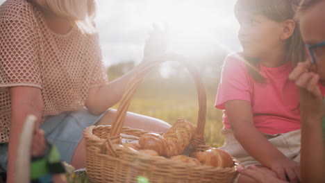 close-up of mother and children seated outdoors for a picnic, woman with a bandaged hand reaches for a snack from a basket as someone passes by