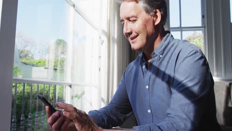 Feliz-Hombre-Caucásico-Mayor-Sentado-En-Una-Soleada-Sala-De-Estar-Usando-Un-Teléfono-Inteligente-Y-Sonriendo