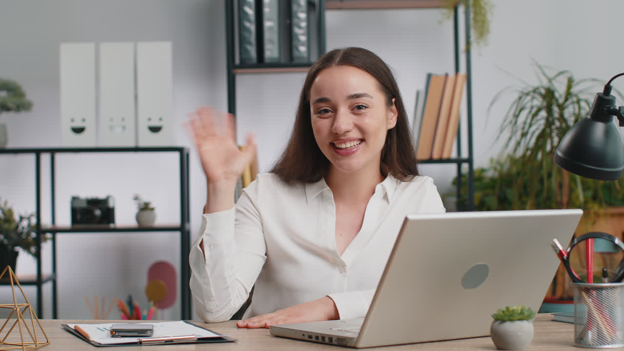 Businesswoman working on laptop smiling friendly at camera and waving hand gesturing hello at office