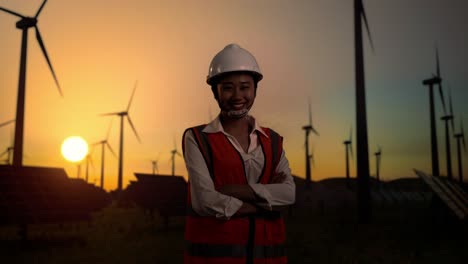 side view of asian female engineer in a helmet smiling and posing with arms crossed while standing in front of wind turbines rotating at sunset