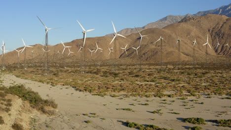 Aerial-view-of-drone-flying-towards-huge-field-of-rotating-wind-turbines-in-the-desert-at-San-Gorgonio-Pass-wind-farm-near-Palm-Springs-Cabazon-in-the-Mojave-desert,-California,-USA