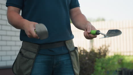 Close up farmer in work apron tossing hand shovels, catching tool then sliding one into pocket on belt at waist, garden background with fence and house, sunny yard