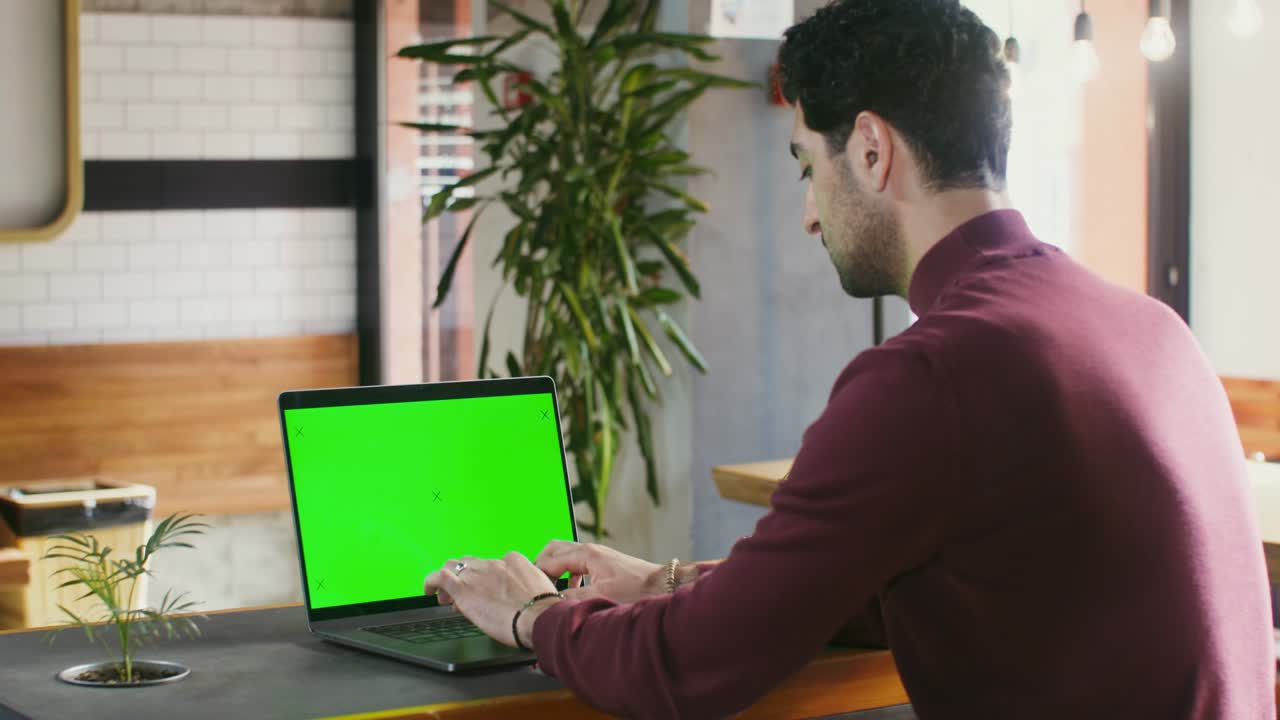 un joven está escribiendo en una computadora portátil con una pantalla verde sentado en una cafetería