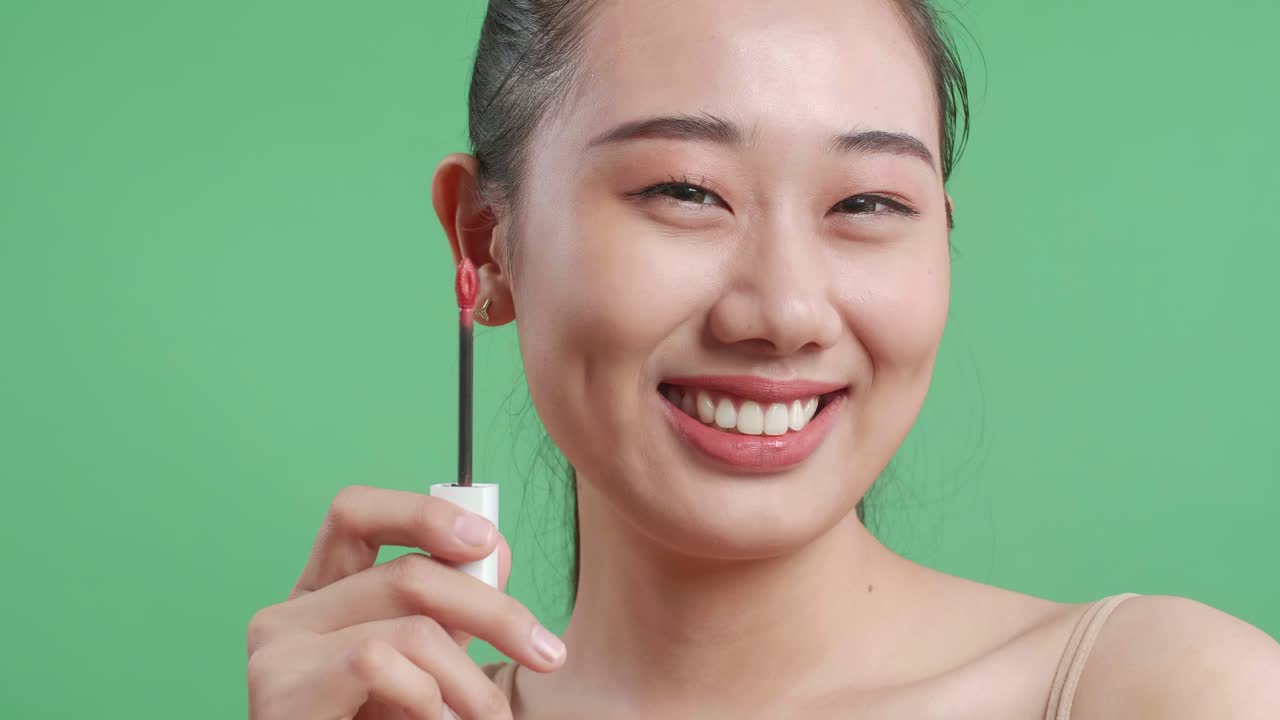 스튜디오에서 녹색 스크린 배경에 서 있는 동안 립스틱을 들고 카메라에 미소 짓는 아시아 여성의 클로즈업