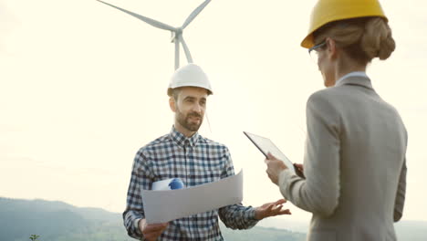 caucasian male engineer wearing a helmet holding blueprints and talking with a female colleage at the huge windmills turbines