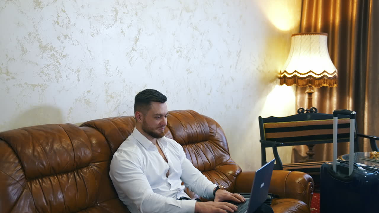 Handsome young businessman works on laptop in a hotel room. Man isresting after businessmeeting and surfing internet.