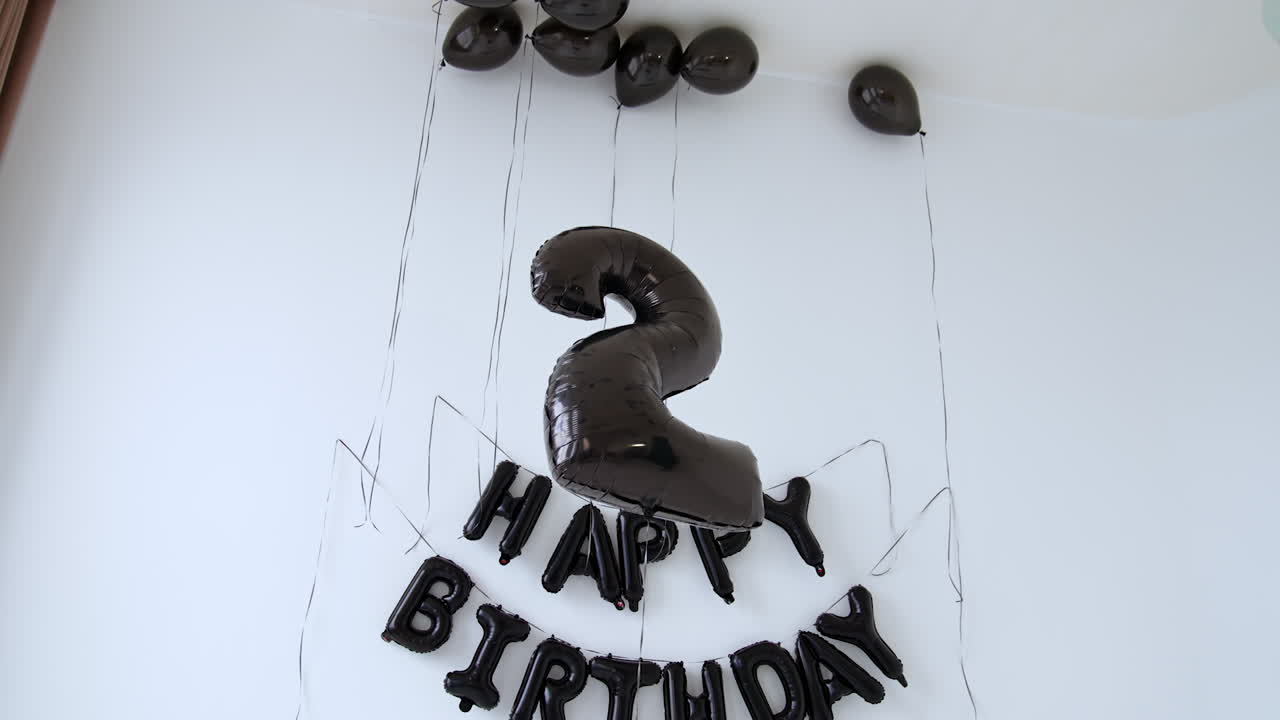 Cute little boy in white shirt, black tie and jeans holding sunglasses and trying to catch a balloon of number two. Happy Birthday sign on the wall at backdrop.