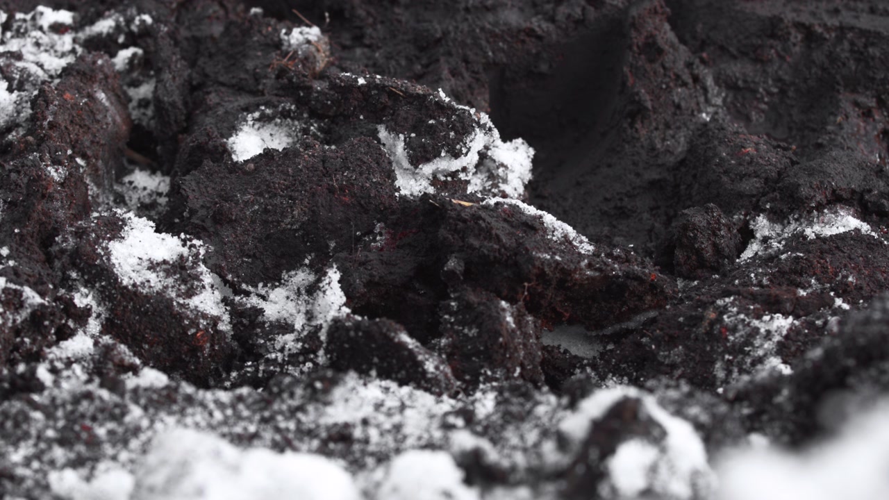 Rugged close-up of dark soil or rock clumps with scattered snow and frost patches, seen from very low viewpoint with shallow depth, muted black, gray, and white tones, textured surface, and cold
